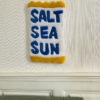 Tapis mural en tufting avec l'inscription 'Salt, Sea, Sun', aux tons bleus et jaunes, fabriqué en laine acrylique avec finition antidérapante.