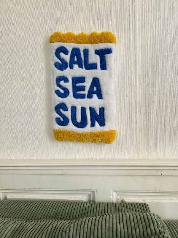 Tapis mural en tufting avec l'inscription 'Salt, Sea, Sun', aux tons bleus et jaunes, fabriqué en laine acrylique avec finition antidérapante.