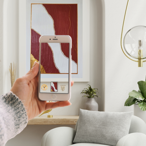 PHOTO 2 etre présent sur Glisstadeco Une personne tenant un téléphone mobile et utilisant la réalité augmentée pour visualiser une décoration murale virtuelle dans son intérieur. L'écran du téléphone montre l'œuvre d'art superposée sur le mur, illustrant comment l'utilisateur peut prévisualiser la décoration avant de l'installer réellement.